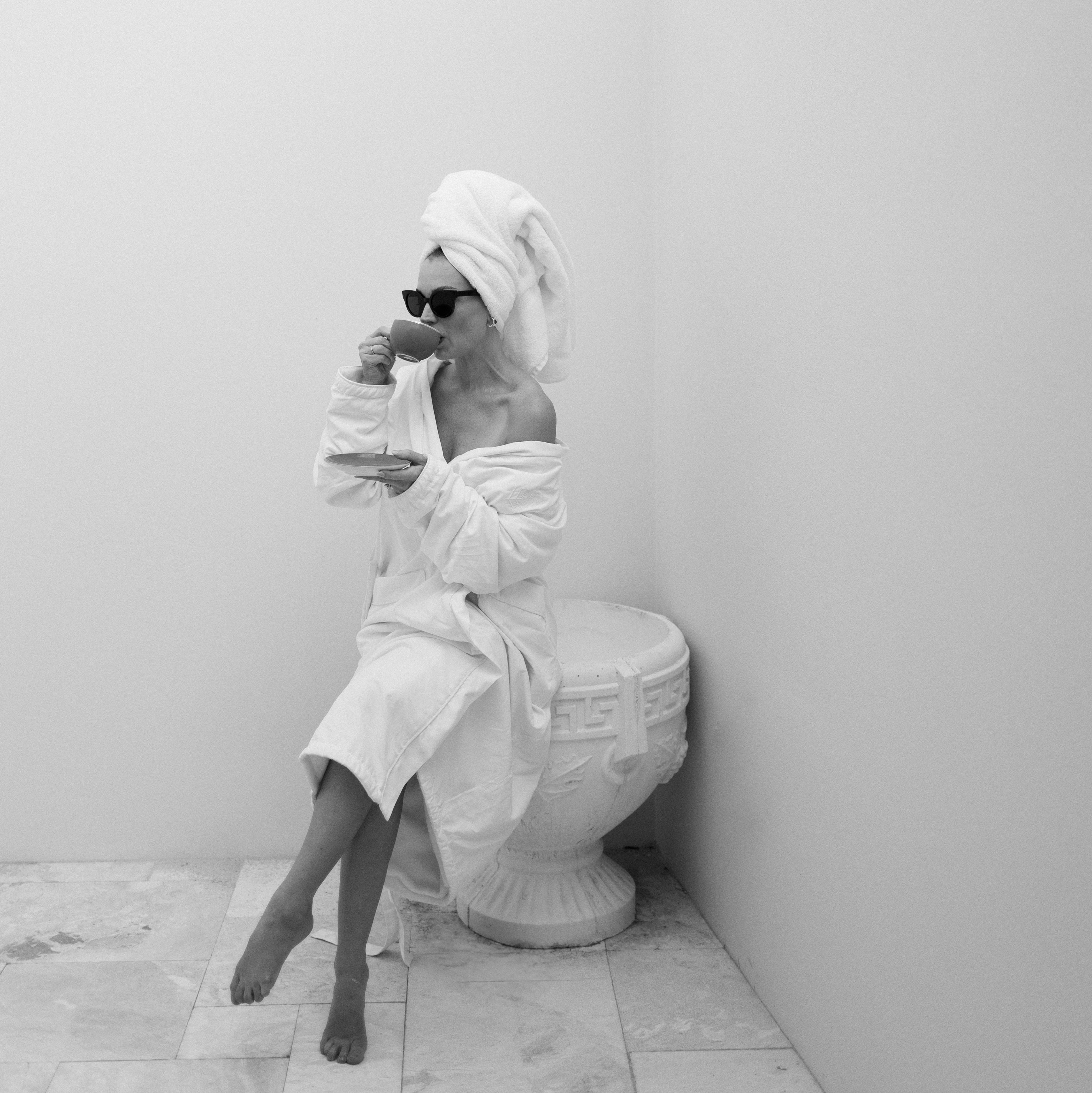 Person sitting on a toilet wearing sunglasses and a robe with a towel wrapped around their head.