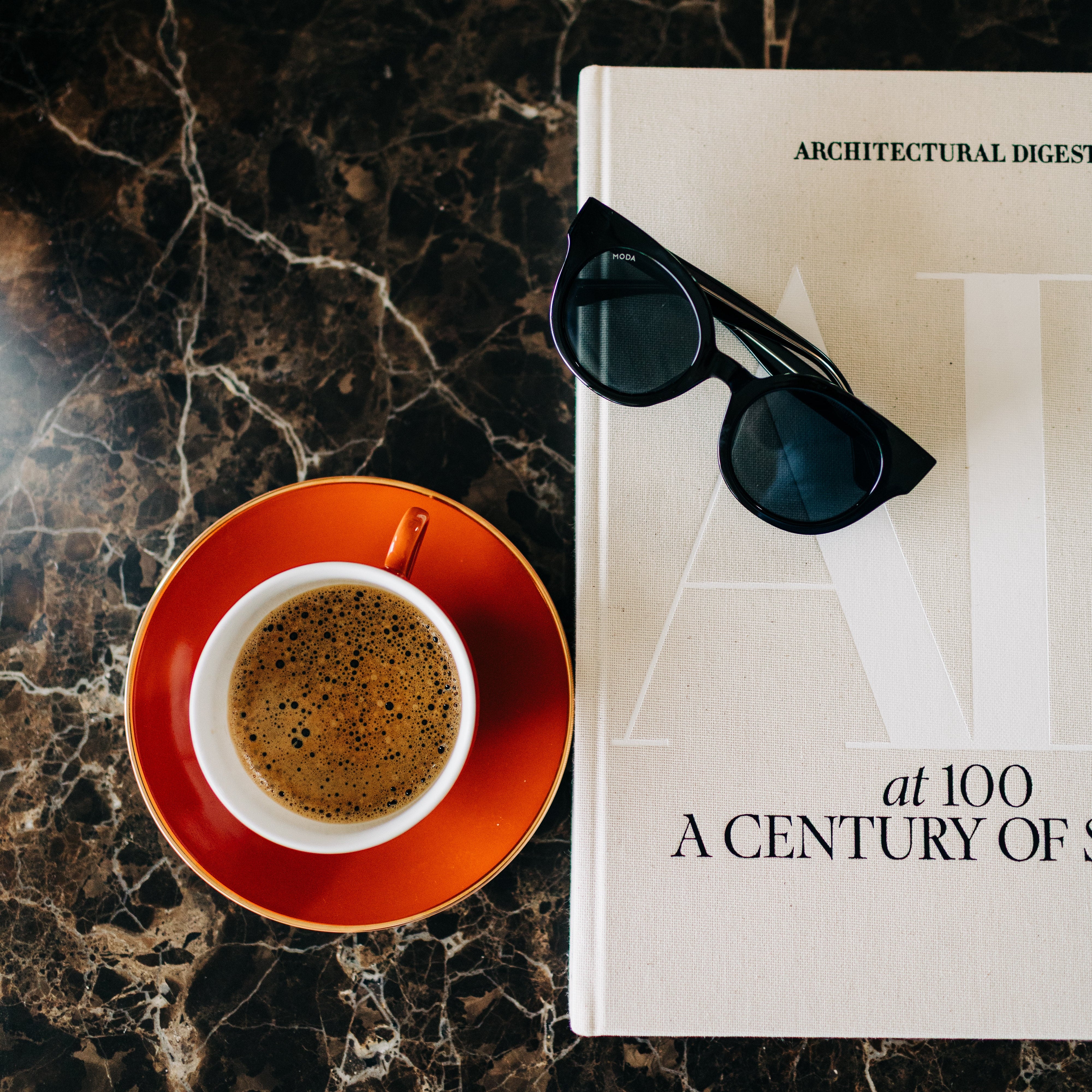 Cup of coffee on a red saucer with sunglasses on top of a magazine on a marble surface